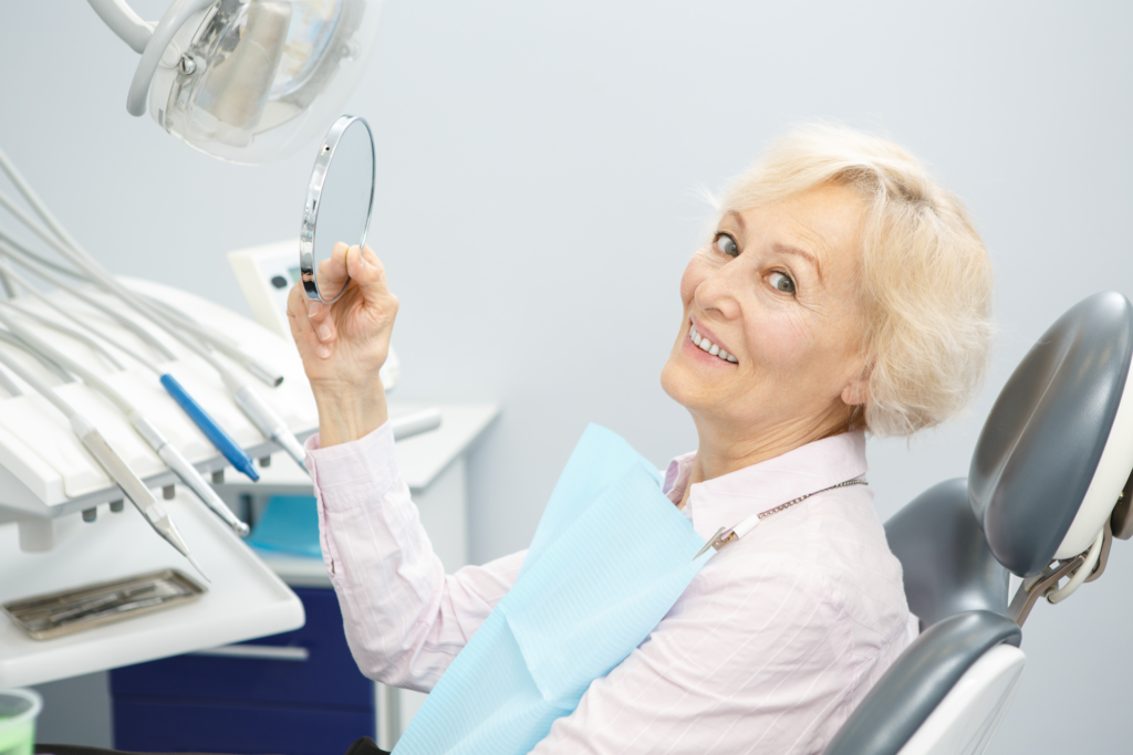 Senior female patient smiling to the camera holding a mirror sitting in a dental chair
